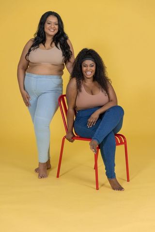 Curvy friends smiling and posing in neutral tanks and jeans on bright yellow backdrop