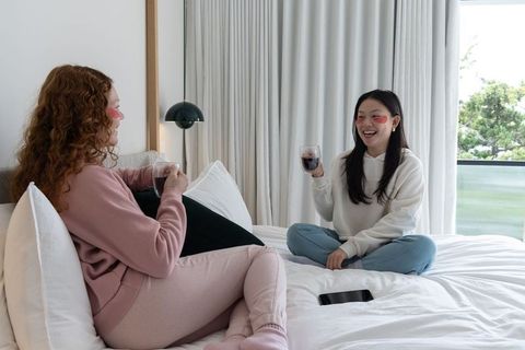Friends relaxing on bed wearing loungewear and hoodies sharing wine and laughter