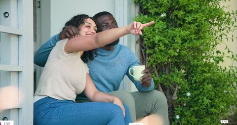 Couple enjoying cozy morning on front porch, pointing out view and sharing coffee