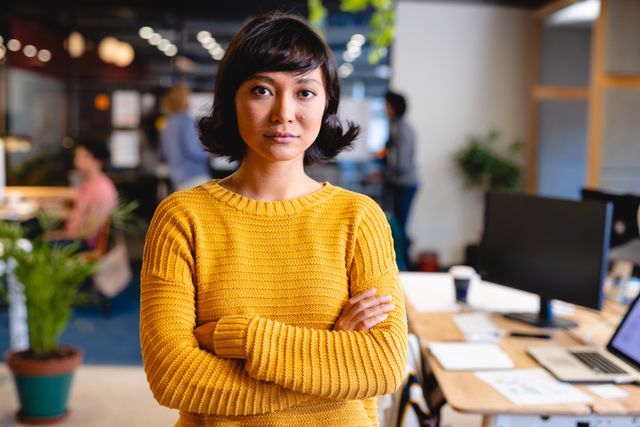 Biracial businesswoman stands confidently with arms crossed in modern creative office space, with colleagues working in background. Suitable for use in promotional materials for entrepreneurship, leadership, teamwork, and modern office culture themes.