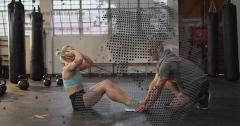 Female athlete performing sit-ups while coach anchoring feet in industrial crossfit gym
