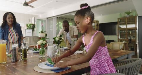 African american family preparing sunlit dining table, girl placing napkin and green leaf
