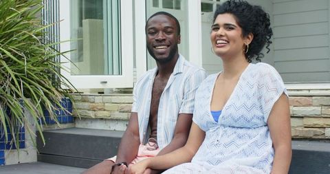 Laughing diverse couple sitting on front porch holding hands, casual suburban companionship