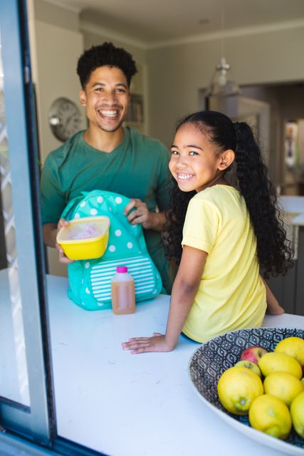 Father packing lunch for daughter in kitchen. Daughter smiling while sitting by counter. Ideal for family lifestyle, parenting tips, morning routine, and healthy eating themes.