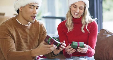 Diverse couple exchanging christmas gifts on sofa wearing santa hats cozy holiday moment