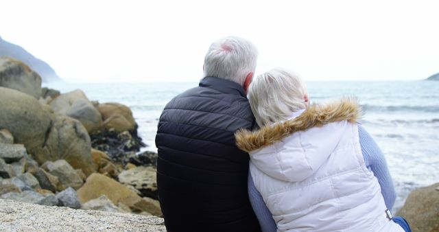 Senior couple sitting on rock looking at ocean. Ideal for topics on aging, retirement living, relationships, couples' activities, and travel. Complementary visual for promoting wellness and mindfulness practices among seniors.