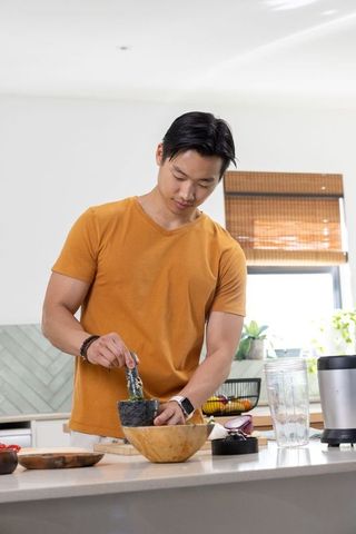 Asian man grinding spices in stone mortar and pestle on kitchen island wearing smartwatch