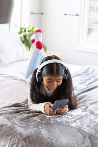 Asian girl lying on bed listening with headphones and using smartphone, cozy bedroom moment