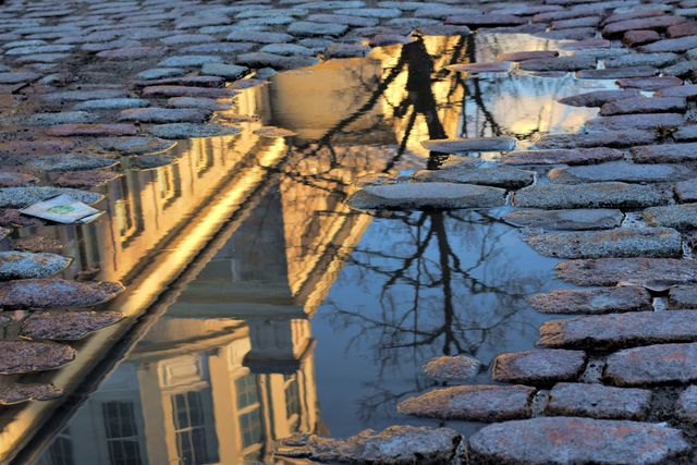 Reflection of a historic building in a water puddle on a cobblestone street. Ideal for concepts of urban tranquility, historic architecture, and atmospheric scenes. Use in articles or promotions related to travel, urban beauty, historical landmarks, or photographic techniques.