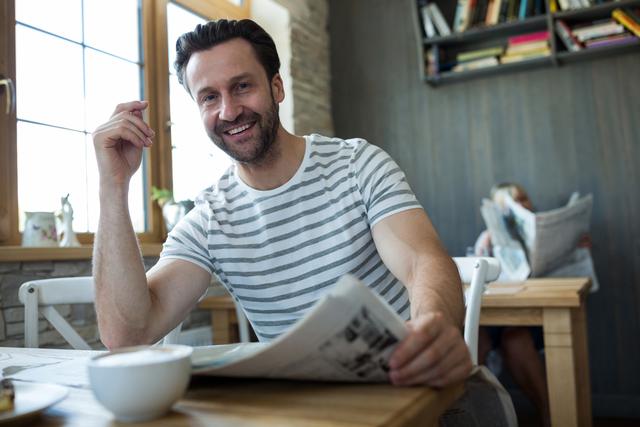 Man enjoying a relaxed morning in a coffee shop, reading a newspaper and smiling. Ideal for use in lifestyle blogs, coffee shop advertisements, or articles about leisure activities and morning routines.