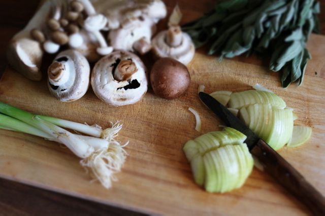 Focused on fresh vegetables and mushrooms being prepared on a wooden cutting board, this image showcases healthy ingredients including onions, green onions, and various mushrooms. This imagery is perfect for use in content related to cooking tutorials, healthy eating, meal preparation, culinary instruction, and organic food recipes. Its rustic kitchen feel makes it suitable for food blogs, recipe books, culinary websites, and health-conscious marketing materials.
