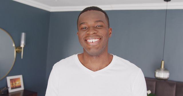Young African American man smiling while standing in well-decorated modern apartment. He appears relaxed and happy. This can be used for lifestyle blogs, interior design features, advertisements, or articles about modern living spaces and personal happiness.