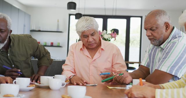This scene shows a group of senior men gathered around a table, engaging in a discussion and writing notes. They appear to be in a relaxed, casual setting with coffee cups and papers in front of them. This image can be used for articles or advertisements focused on senior activities, the importance of social interactions in elderly communities, or teamwork among mature adults.