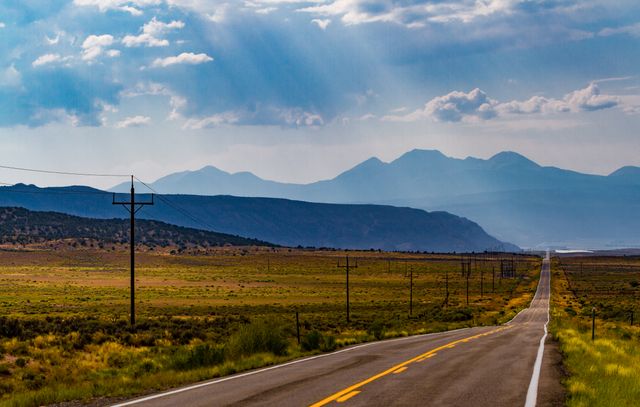 Image showcases a long straight road cutting through open countryside with mountains in the background. Ideal for use in travel blogs, adventure websites, and advertisements promoting road trips. Use it to highlight themes of solitude, travel, and open spaces.