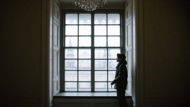 Contemplative man gazing through tall sash window in historic interior, chandelier glow