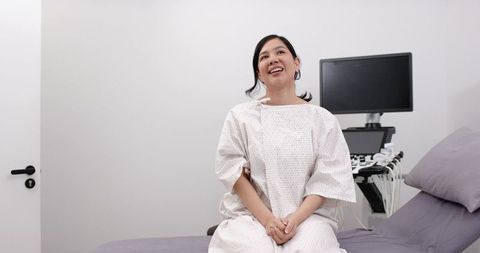 Asian woman sitting on exam bed wearing medical gown beside ultrasound machine smiling