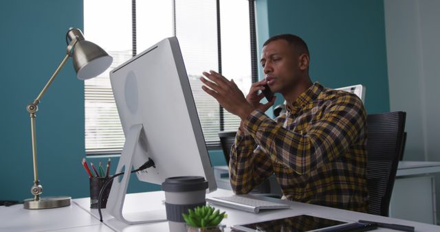 Young professional making a phone call while working on computer in a modern office. Ideal for content about business, problem solving, technology in the workplace, office communication, and multitasking skills.