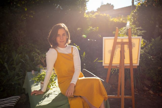 Caucasian woman in yellow dress posing in a garden with a canvas and easel, enjoying free time alone. Sunlight filters through the trees, creating a serene and creative atmosphere. Ideal for use in lifestyle, art, and relaxation themes, promoting outdoor activities, creativity, and peaceful moments at home.