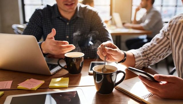 Two coworkers collaborating over coffee in sunlit coworking space using laptop smartphone