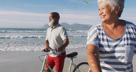 Smiling mature couple strolling with bicycles on sunlit beach with gold confetti and ocean