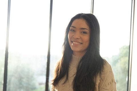 Smiling woman standing by window wearing cream knit sweater, sunlit rim-lit portrait