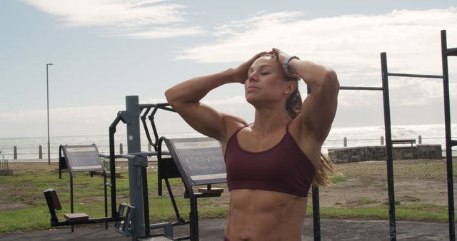 Muscular woman resting with hands on head after an intense workout at an outdoor gym by the sea. The image conveys strength, determination, and focus on fitness near a beautiful beach setting. Ideal for promoting fitness regimens, active lifestyle brands, health and wellness programs, and motivational fitness content.