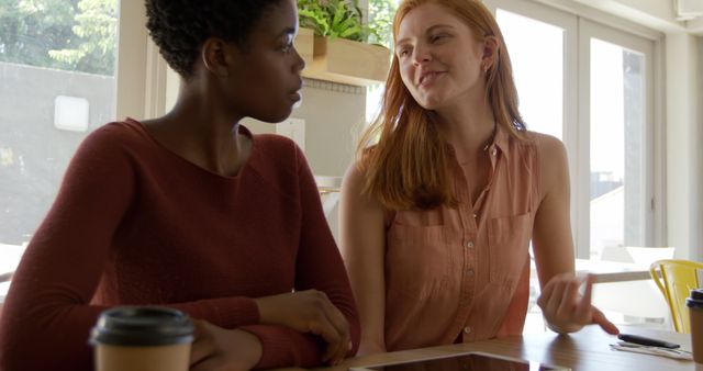 Two women are sitting together at a coffee shop, engaged in a cheerful conversation with a digital tablet in front of them. Ideal for illustrations of casual business meetings, teamwork, collaboration, and social interaction in a relaxed, modern setting. This image can be used for articles on coworking spaces, remote work, or social gatherings.