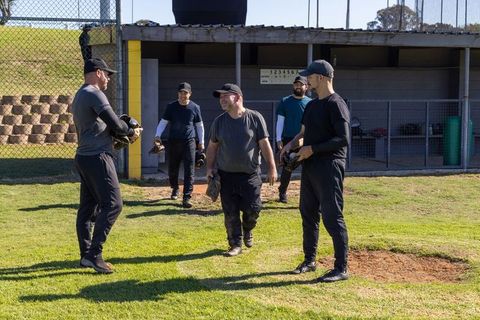Five male baseball players discussing play near dugout during sunny infield practice