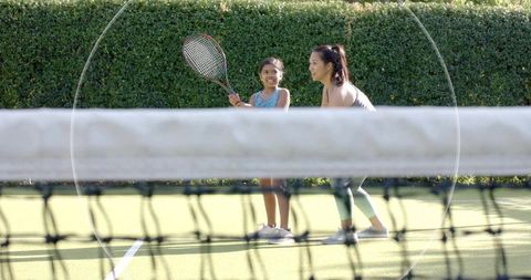 Woman coaching young girl in tennis on sunlit outdoor turf court during private lesson
