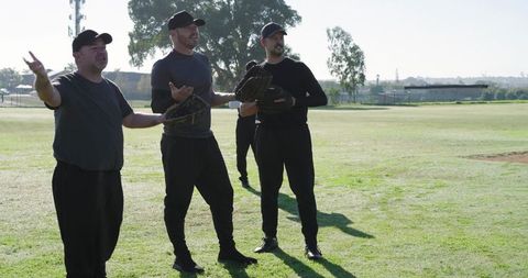 Mature teammates standing on sunlit baseball field holding gloves and caps during practice