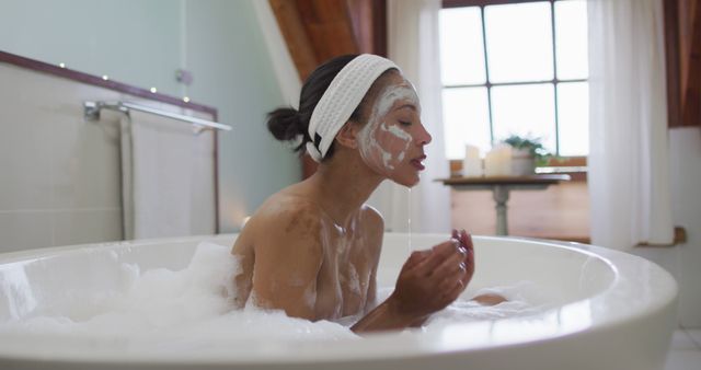 Biracial woman sitting in bathtub taking a bath, cleaning her face. domestic life, spending quality free time relaxing at home.
