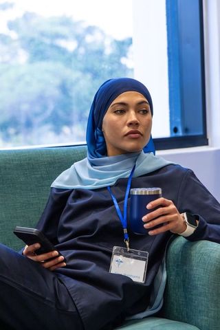 Professional woman in blue headscarf relaxing in office lounge with coffee and smartphone