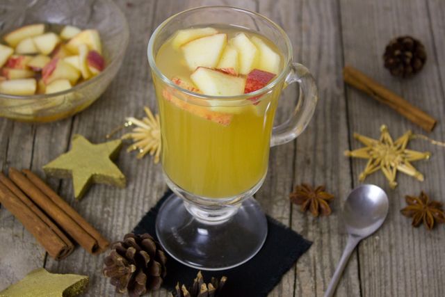 Appealing visual of a hot apple cider drink garnished with apple chunks, served in a glass mug on a rustic wooden table. Surrounded by cinnamon sticks, star anise, pinecones, and festive decorations, this image is ideal for use in holiday recipes, social media posts about fall or winter festivals, or articles related to cozy home aesthetics and seasonal beverages.