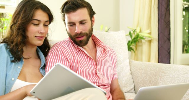 Couple comfortably seated on couch using tablet and laptop. Learn new technological skills together, browse online content or plan activities. Ideal for depicting modern lifestyle, remote work, online shopping or digital communication themes.