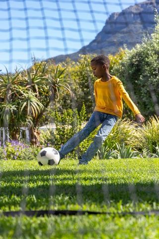 African american boy kicking soccer ball in sunlit backyard garden with mountain backdrop