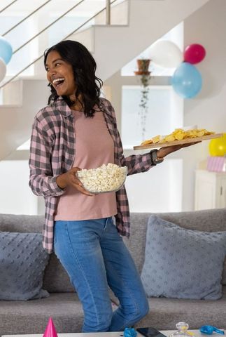 Laughing woman holding popcorn bowl and chips platter at colorful home party