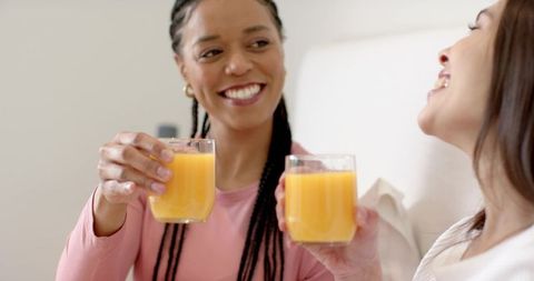Clinking glasses of orange juice on couch smiling friends enjoying relaxed morning