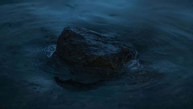 Dark wet rock rising from calm lake at dusk creating concentric ripples and moody twilight