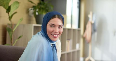 Indian woman smiling while wearing blue hijab and sitting on grey sofa in modern lounge