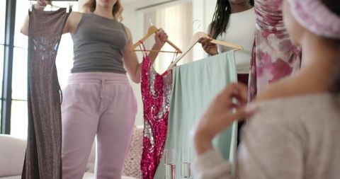 Female friends holding dresses showing options to seated girl during at-home dress fitting