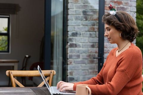 Woman typing on laptop at outdoor terrace table wearing festive headband and burnt orange sweater
