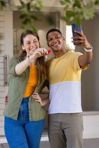 Young diverse couple celebrating new home while taking selfie and holding house keys