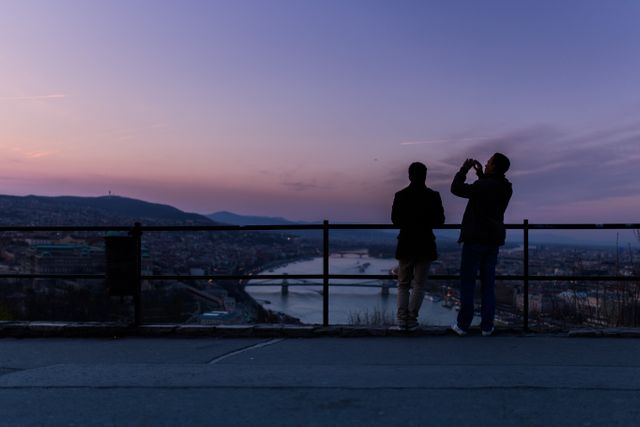Two individuals are standing by a railing, observing a city skyline during a stunning sunset. One person is taking a photo. Great for tourism, travel, urban lifestyle, and skyline photography themes.