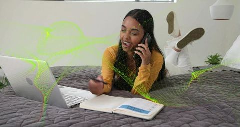 Young woman studying and talking on phone with laptop on bed, multitasking comfortably