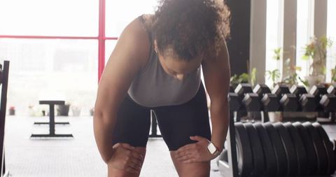 Female athlete bending forward catching breath after strength training by dumbbell rack