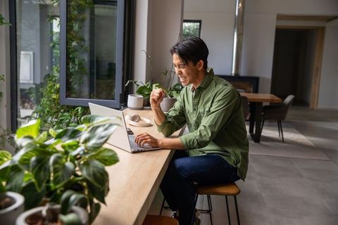 Asian man cheering while working remotely typing on laptop at sunlit plant-filled home workspace