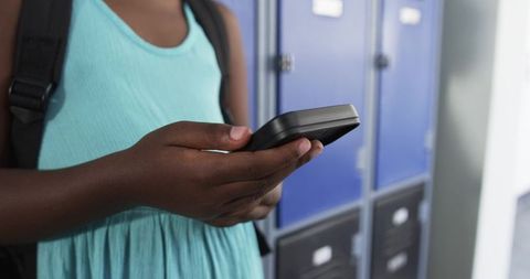 African student texting by blue lockers in school hallway wearing teal dress and backpack