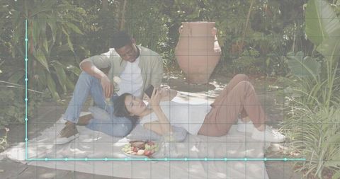 Couple relaxing on blanket in secluded sunlit garden sharing phone and fresh fruit