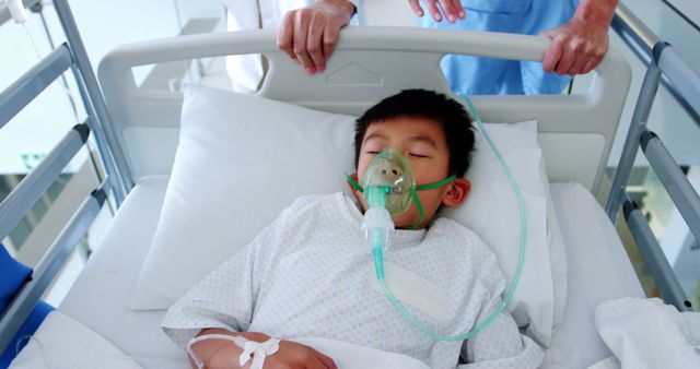 A young Asian boy is receiving medical care in a hospital bed, with a healthcare professional by his side, with copy space. His condition necessitates the use of an oxygen mask, indicating a serious respiratory issue or surgery recovery.