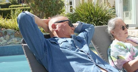 Senior couple relaxing on poolside lounge chairs wearing sunglasses in sunny backyard